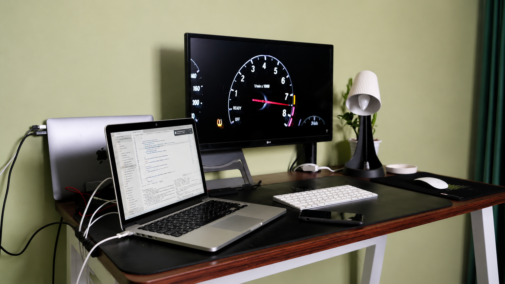 An earlier desk: an LG monitor, an open MacBook Pro showing code, a Magic Keyboard and Magic Mouse, a lamp and a plant